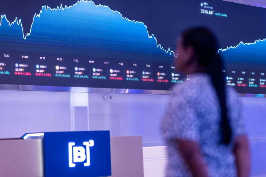 Painel de índices financeiros em queda na bolsa de valores B3; pessoa desfocada ao fundo observando o gráfico.