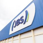 Fachada de prédio industrial da JBS com logotipo em destaque, refletindo estrutura da empresa no setor alimentício.