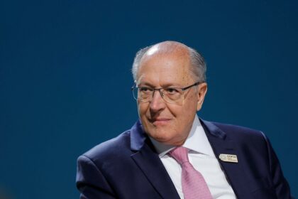 Homem de terno escuro e gravata rosa em ambiente formal, usando óculos, com fundo azul.