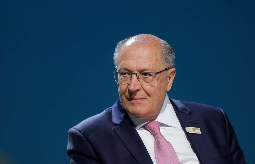 Homem de terno escuro e gravata rosa em ambiente formal, usando óculos, com fundo azul.
