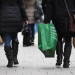 Duas pessoas vestindo casacos e botas caminham carregando sacolas de compras em uma rua movimentada.
