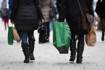 Duas pessoas vestindo casacos e botas caminham carregando sacolas de compras em uma rua movimentada.
