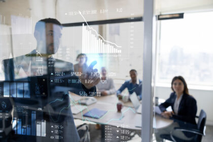Homem interage com gráficos digitais em sala de reuniões, colegas ao fundo. Análise de dados empresariais em destaque.