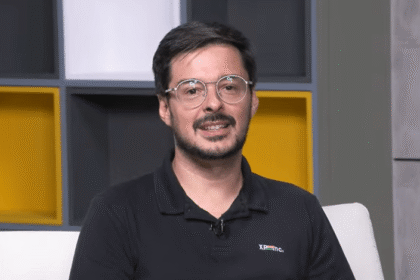 Homem de óculos fala em ambiente interno moderno, com estantes coloridas ao fundo, transmitindo profissionalismo e tecnologia.