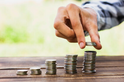 Mão empilhando moedas sobre mesa de madeira ao ar livre, representando crescimento financeiro e economia.