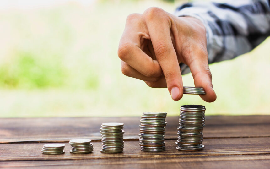 Mão empilhando moedas sobre mesa de madeira ao ar livre, representando crescimento financeiro e economia.