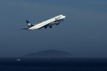 Avião da Azul decola sobre o oceano, com ilhas ao fundo, destacando-se no céu azul claro.