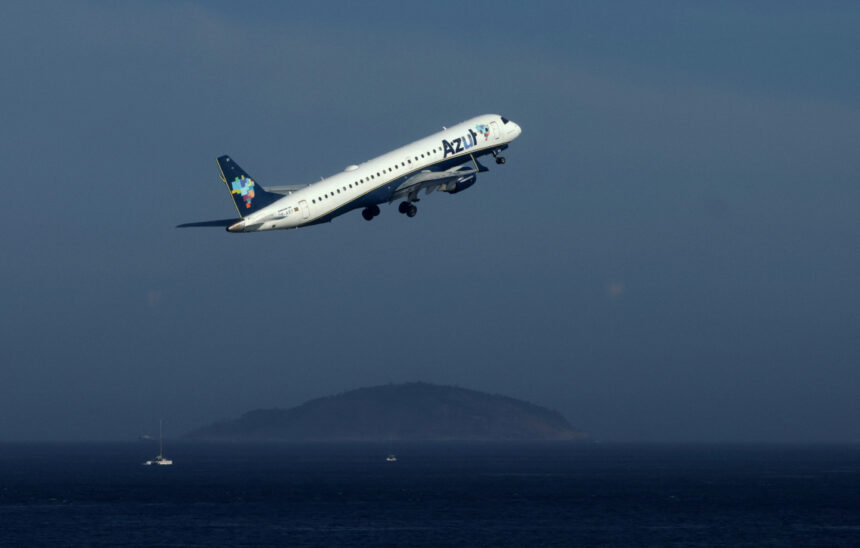 Avião da Azul decola sobre o oceano, com ilhas ao fundo, destacando-se no céu azul claro.