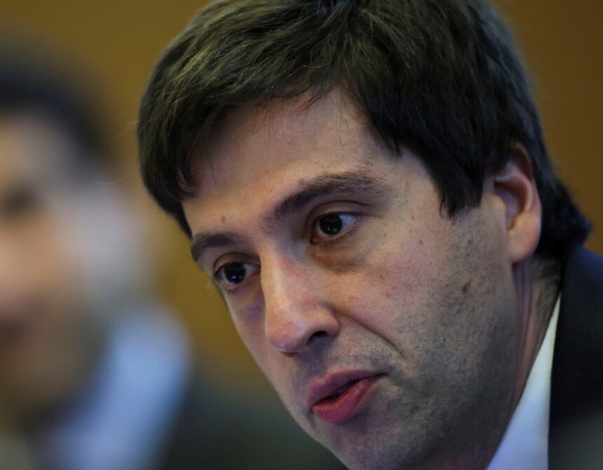 Homem de terno azul em close-up durante evento, exibindo expressão séria e pensativa; fundo desfocado.