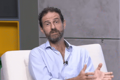 Homem de camisa azul conversando enquanto gesticula, sentado em cadeira branca em ambiente de escritório.