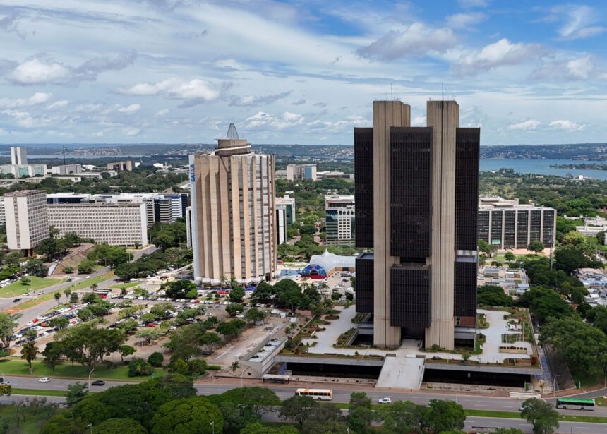 Paisagem urbana de Brasília com edifícios oficiais, árvores ao redor e ao fundo o Lago Paranoá sob céu parcialmente nublado.