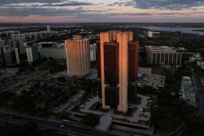 Vista aérea de Brasília ao entardecer, destacando grandes edifícios e vias movimentadas, com o Lago Paranoá ao fundo.