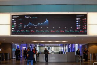 Painel de cotações da bolsa de valores em um saguão movimentado, exibindo gráfico e índices financeiros. Pessoas caminham ao redor.