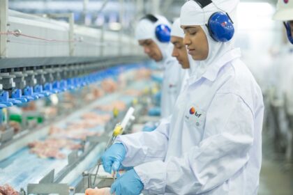 Trabalhadores em linha de produção de alimentos, usando trajes de proteção e fones, inspecionam produtos em esteira automatizada.