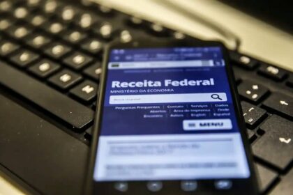Celular acessa site da Receita Federal sobre teclado de computador, destacando serviços online do governo.