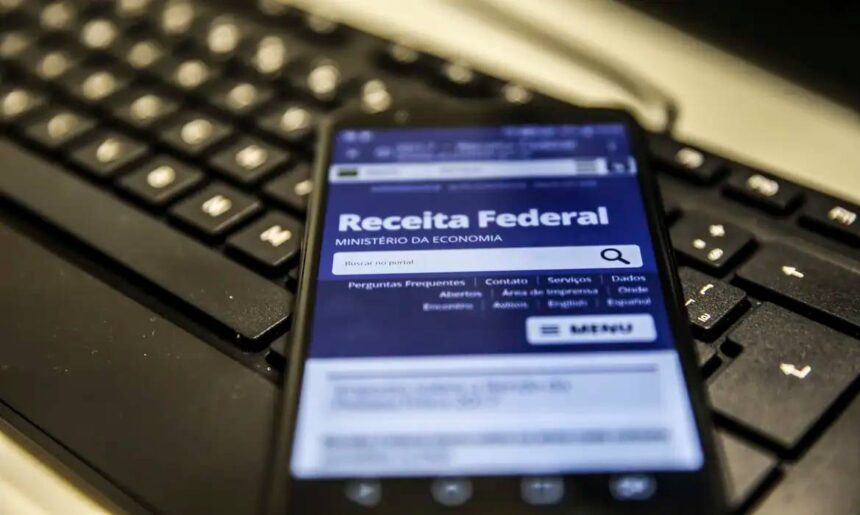 Celular acessa site da Receita Federal sobre teclado de computador, destacando serviços online do governo.