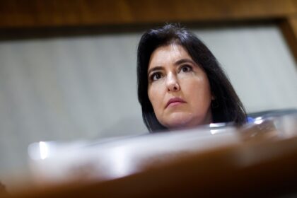 Mulher com expressão séria em ambiente interno, olhando para cima, com parte do rosto iluminada.