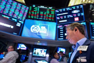 Corretor na bolsa de valores de Nova York com vários monitores exibindo cotações e gráficos, em ambiente de negociação agitado.