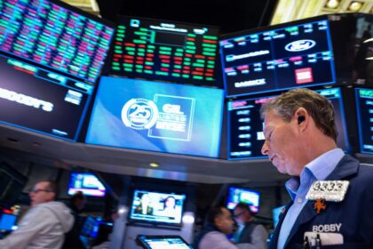 Corretor na bolsa de valores de Nova York com vários monitores exibindo cotações e gráficos, em ambiente de negociação agitado.