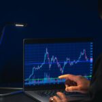 Pessoa analisando gráficos de ações em laptop sob baixa iluminação, destacando tendências do mercado financeiro.