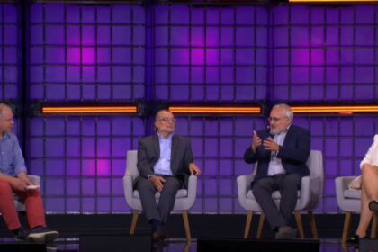 Quatro pessoas em um painel de discussão, um homem fala gesticulando, enquanto outras três pessoas ouvem atentamente. Fundo roxo.