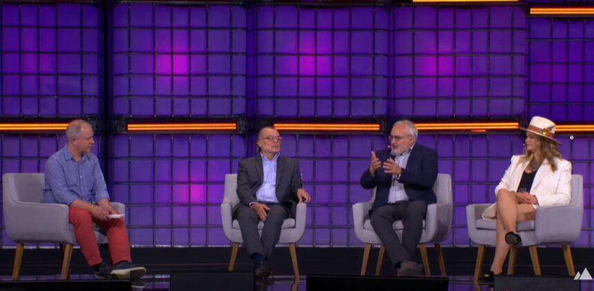 Quatro pessoas em um painel de discussão, um homem fala gesticulando, enquanto outras três pessoas ouvem atentamente. Fundo roxo.