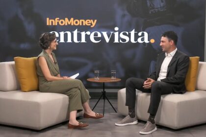 Duas pessoas sentadas em um estúdio discutindo questões financeiras em um programa de entrevista com fundo temático e copos de água.