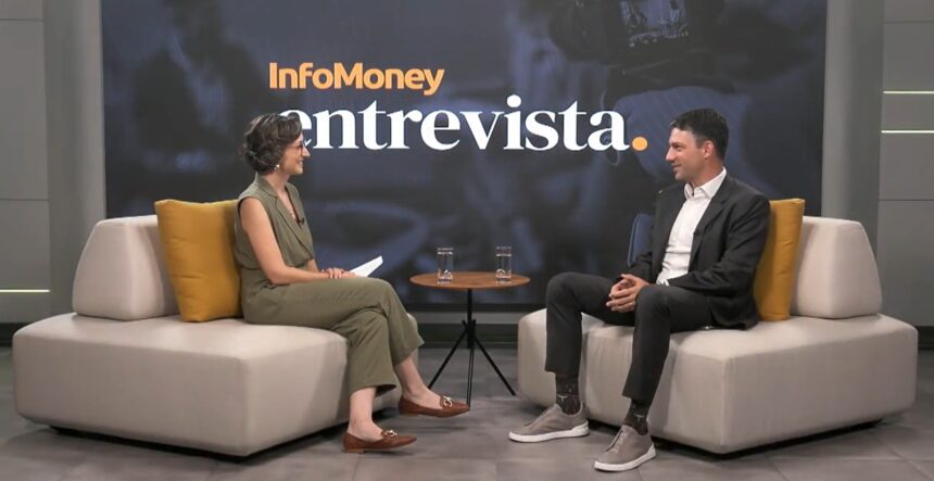 Duas pessoas sentadas em um estúdio discutindo questões financeiras em um programa de entrevista com fundo temático e copos de água.