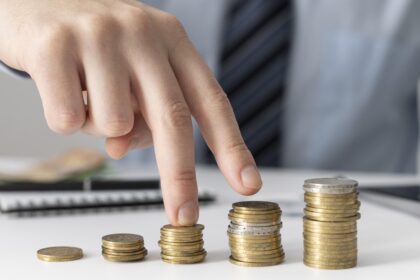 Uma mão contando quatro pilhas crescentes de moedas, representando planejamento financeiro e economia.