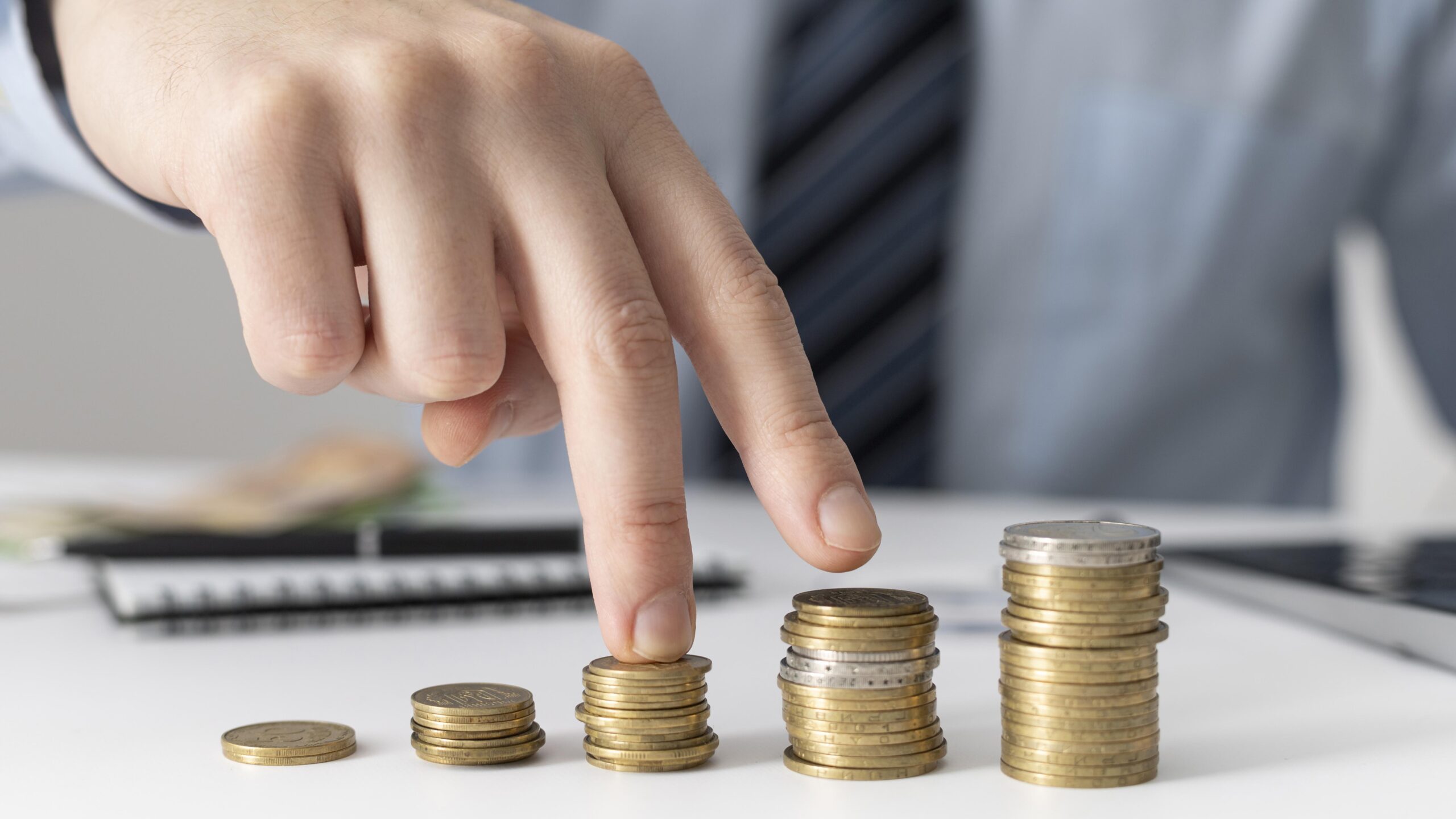 Uma mão contando quatro pilhas crescentes de moedas, representando planejamento financeiro e economia.