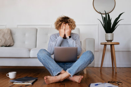 Homem frustrado sentado no chão, mãos no rosto, com notebook no colo. Ambiente doméstico com sofá, planta e caderno ao redor.