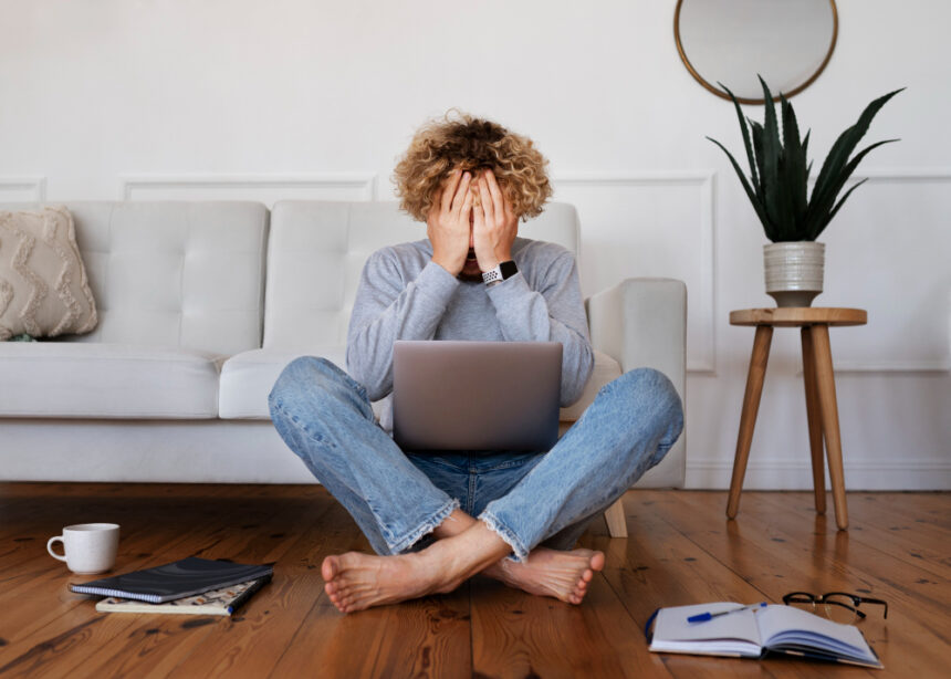 Homem frustrado sentado no chão, mãos no rosto, com notebook no colo. Ambiente doméstico com sofá, planta e caderno ao redor.