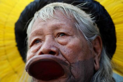 Ancião indígena com cocar amarelo tradicional e disco labial, simbolizando a cultura e tradição de sua tribo.
