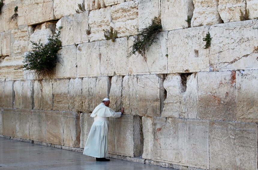 Indivíduo em trajes brancos reza no Muro das Lamentações, em Jerusalém, um local sagrado e histórico de peregrinação.