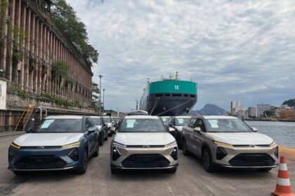Carros elétricos aguardam embarque em um porto com grande navio ao fundo, destacando transporte sustentável e tecnologia.