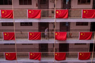 Corredor de edifício com bandeiras da China penduradas, mostrando patriotismo. Pessoa passando ao fundo, usando máscara.