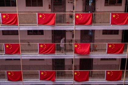 Corredor de edifício com bandeiras da China penduradas, mostrando patriotismo. Pessoa passando ao fundo, usando máscara.