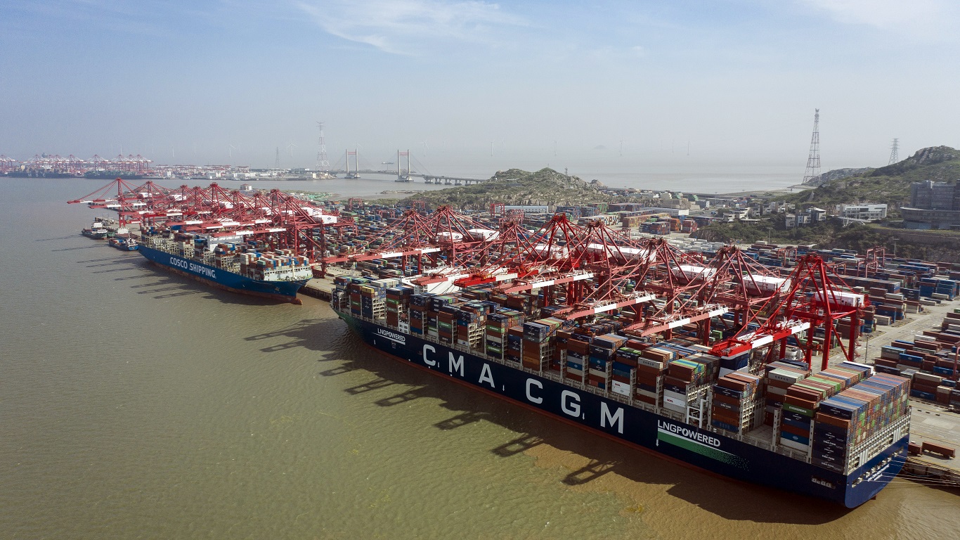Navios de carga atracados em um movimentado porto, com guindastes vermelhos operando contêineres. Paisagem portuária ao fundo.
