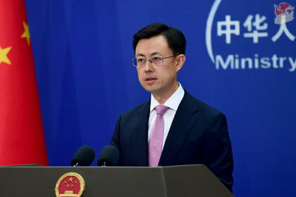 Político em conferência de imprensa em Pequim, com bandeira da China ao lado, discursando de terno e gravata.