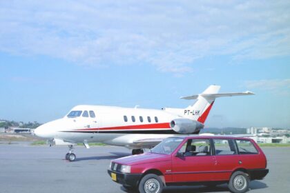 Avião jatinho na pista de decolagem, com carro vermelho ao lado em aeroporto, céu azul ao fundo.