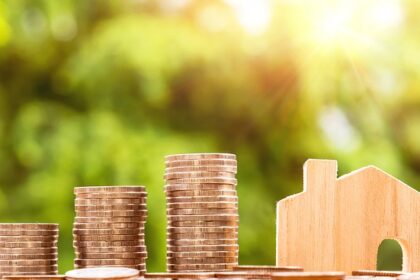 Moedas empilhadas ao lado de uma casa em miniatura, simbolizando o conceito de economia doméstica e investimento em imóveis.