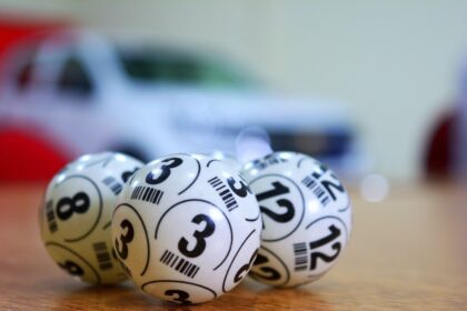 Bolas de bingo numeradas em primeiro plano, destacando os números 3 e 12, com um carro desfocado ao fundo.