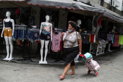 Mulher caminha em uma rua comercial puxando um carrinho de compras, passando por manequins com roupas de verão.