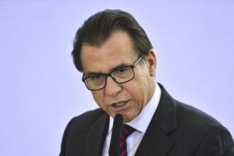 Homem de óculos e terno fala ao microfone em evento formal, transmitindo seriedade e profissionalismo.