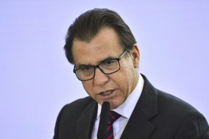 Homem de óculos e terno fala ao microfone em evento formal, transmitindo seriedade e profissionalismo.