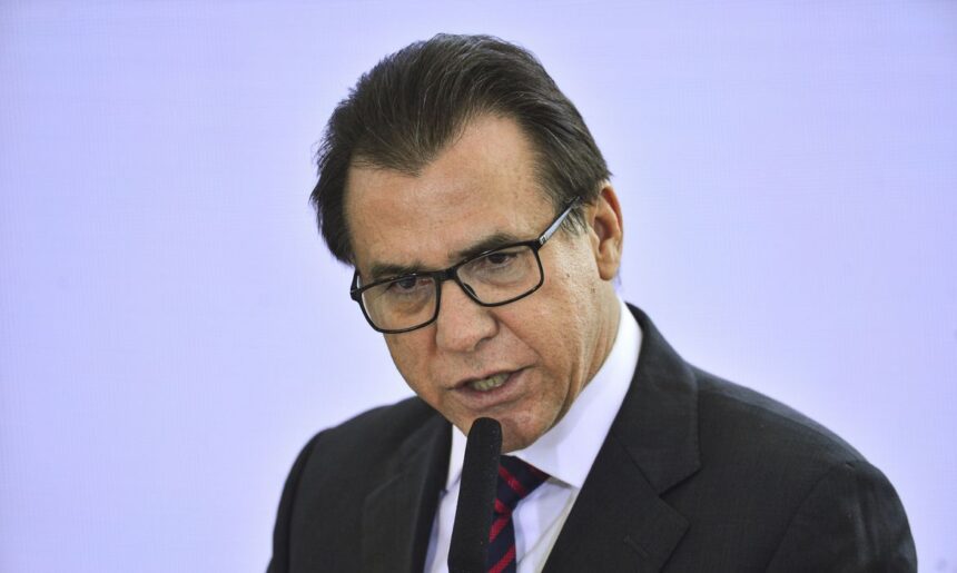 Homem de óculos e terno fala ao microfone em evento formal, transmitindo seriedade e profissionalismo.