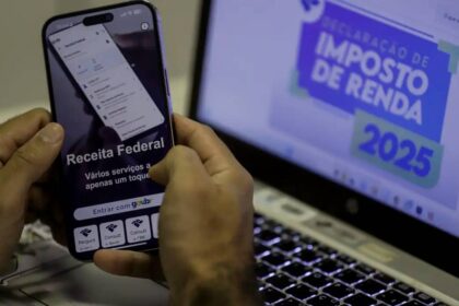 Pessoa utilizando aplicativo da Receita Federal em smartphone, com tela de notebook exibindo Declaração de Imposto de Renda 2025.
