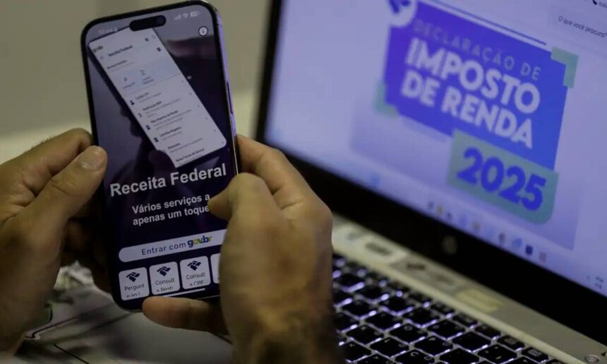 Pessoa utilizando aplicativo da Receita Federal em smartphone, com tela de notebook exibindo Declaração de Imposto de Renda 2025.