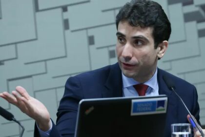 Homem em traje formal gesticula durante discurso em uma sessão no Senado Federal, com microfone e laptop à frente.