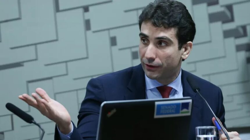 Homem em traje formal gesticula durante discurso em uma sessão no Senado Federal, com microfone e laptop à frente.